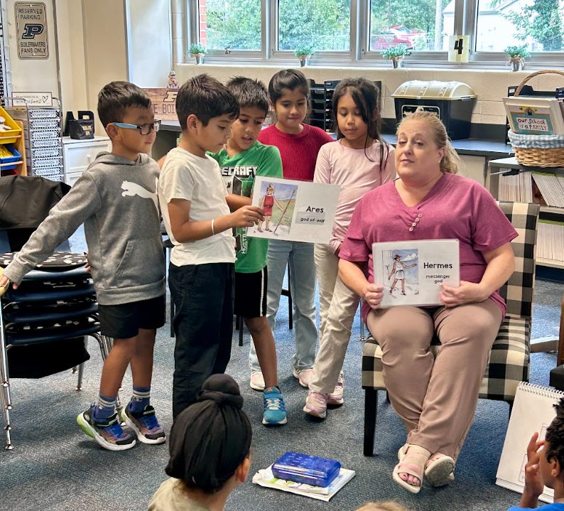 Ms. Goodman holding up a sign with a greek god info with students standing up around and behind her holding a different greek god info card
