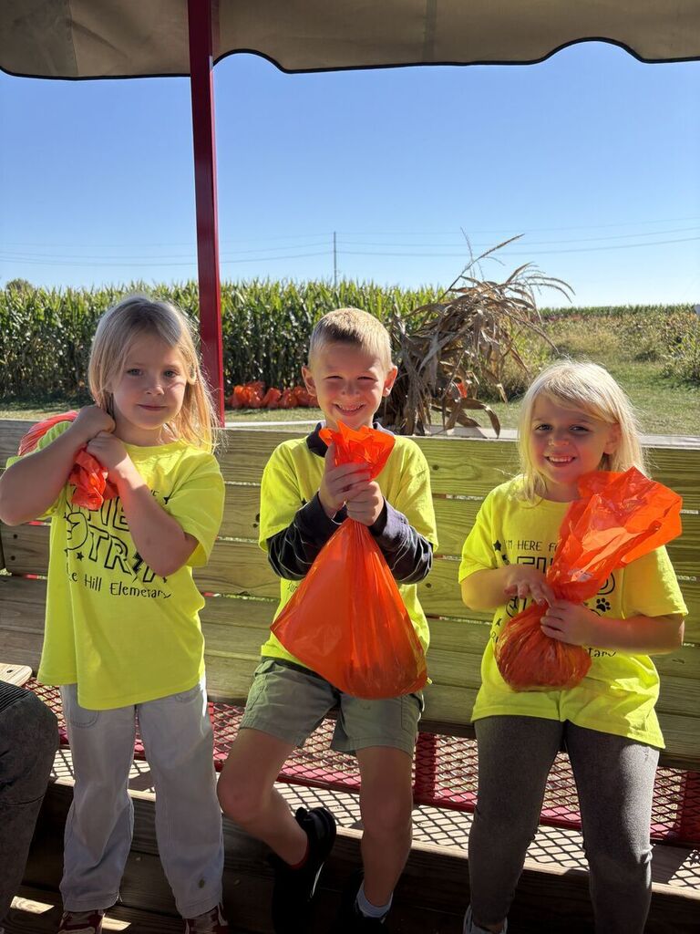 three students sitting on metal bench on hayride smiling with orange bags 