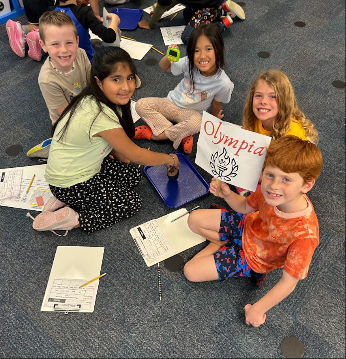 students smiling in a group sitting on the floor holding up their team name sign that says "OLYMPIA"