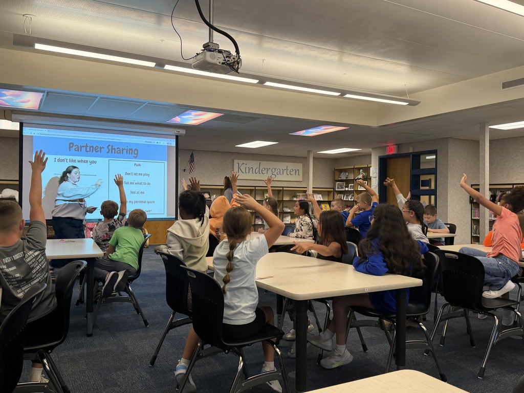students seated at tables raising their hands while teacher is up at the projector screen calling on someone