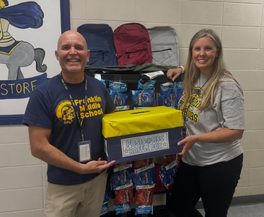 Two adults, one in a blue shirt, the other in a gray shirt, hold a donation box.