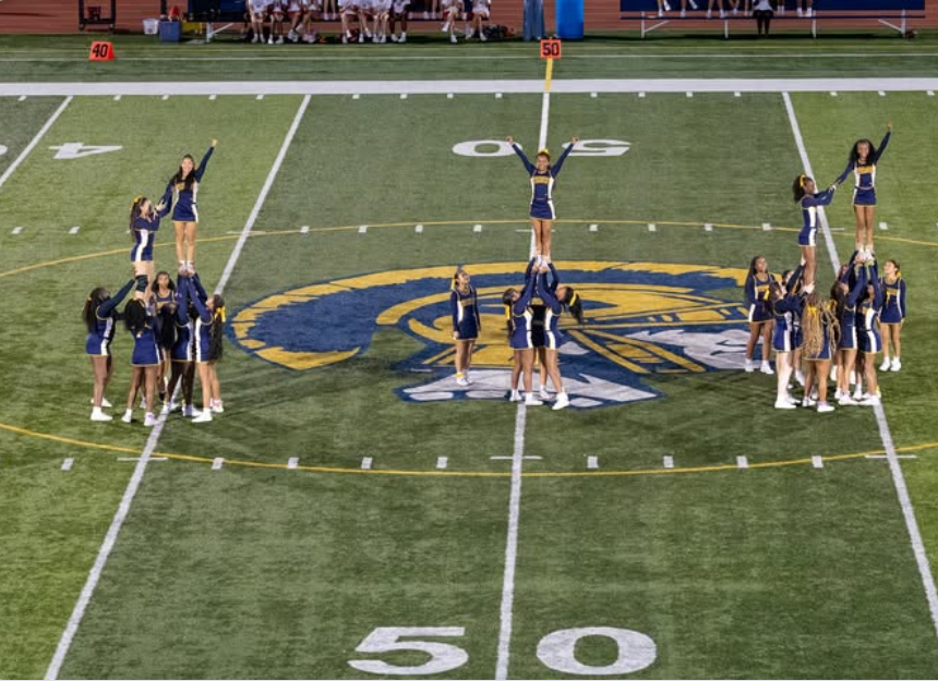 Group of cheerleaders on a football field performing stunts; school logo at center.