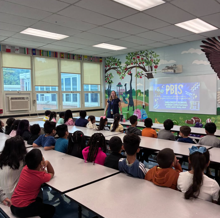 Classroom with painted walls; a teacher stands in front of a projector screen. Students seated at tables listen.