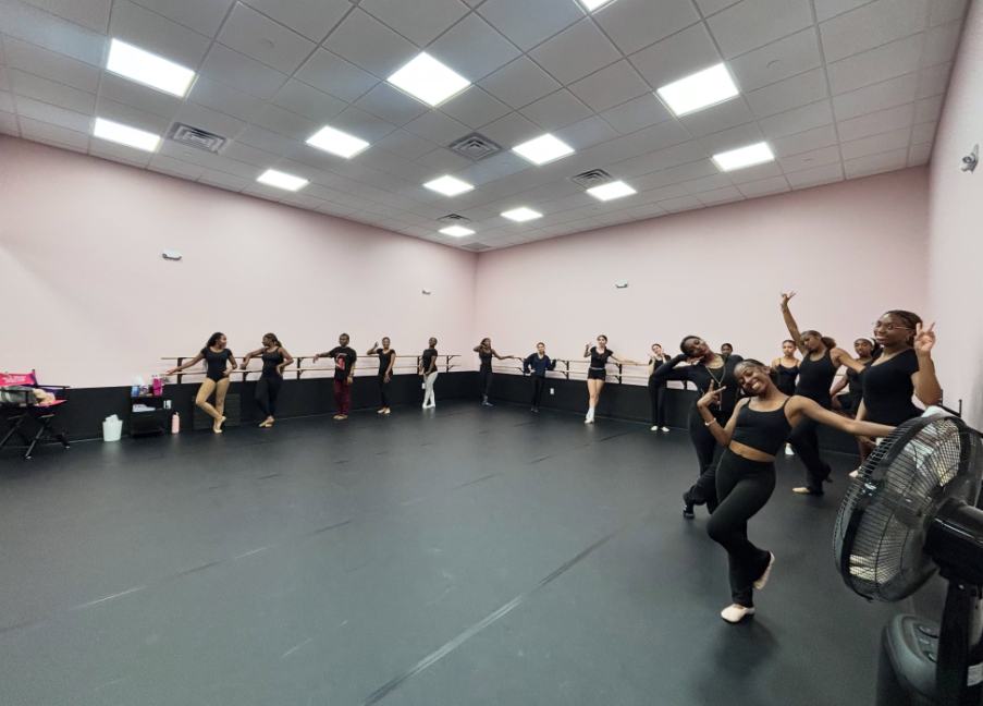 A studio with pink walls and black floors. A group of people practice dance moves, wearing black outfits.