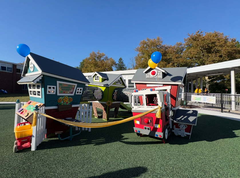 Colorful play structures with balloons on top and a fire truck. Background includes a school building and trees.
