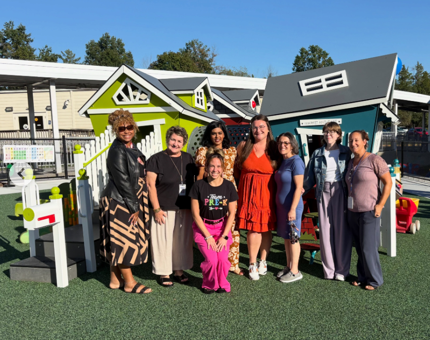 Group of people standing in front of colorful playhouses on a playground, with a fence and trees in the background.