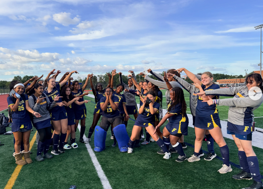 Group of female athletes in blue uniforms on a field celebrating. A blue sky with clouds in the background.