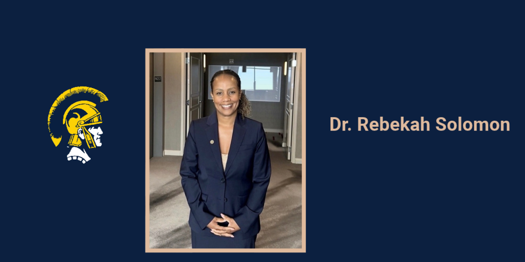 Blue background with a logo. A woman in a navy suit stands in a doorway, smiling and hands clasped.