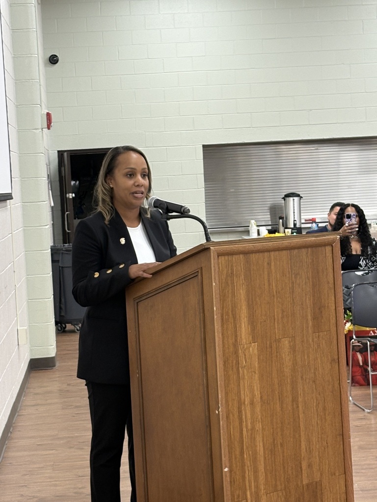 A woman stands at a podium with a microphone, speaking to a room with people seated in chairs.