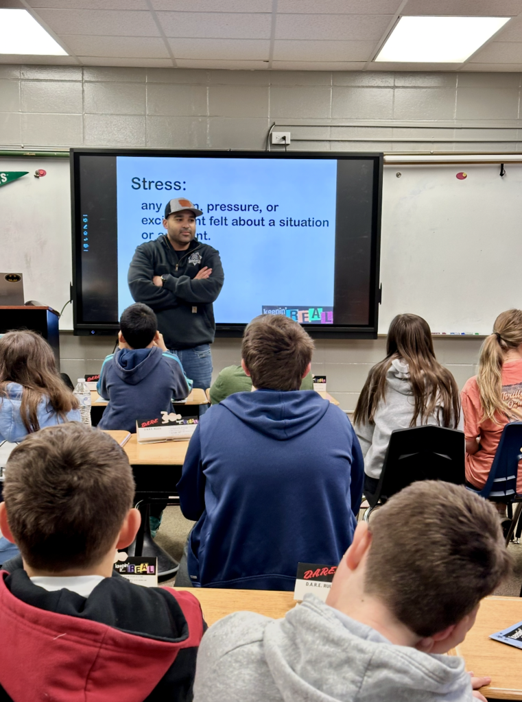 Sarget Ashton Luke standing in the front of a classroom with students listening to him talk. The topic on the screen behind him is stress.