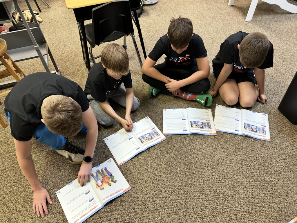 Four boys sit on a carpeted floor, each focused on an open workbook in front of them, in a classroom setting.