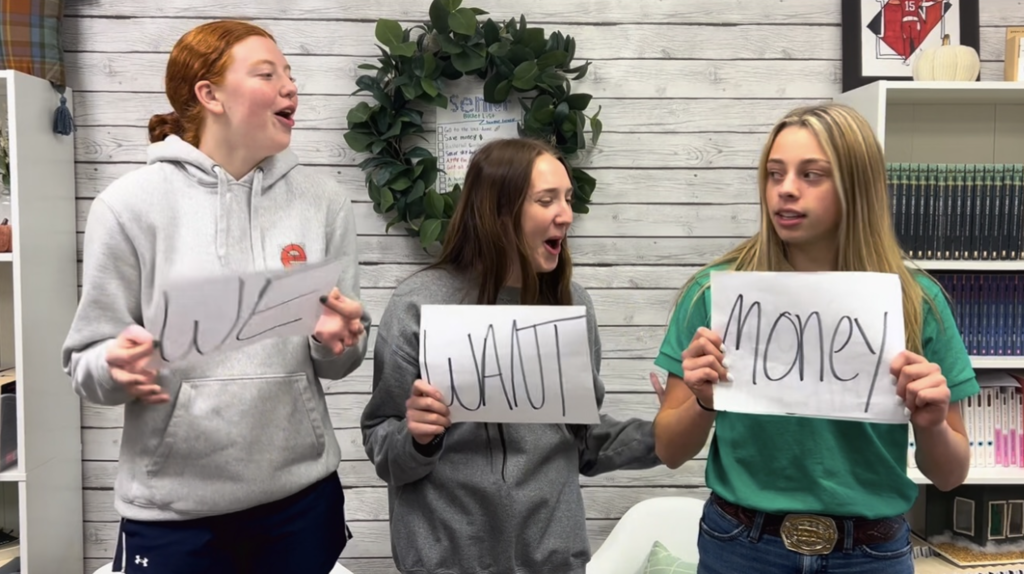 three students holding signs that say "We want money" for video