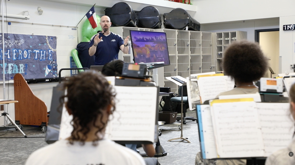 Band conductor standing with hands raised to lead a rehearsal as students view music sheets in the foreground.