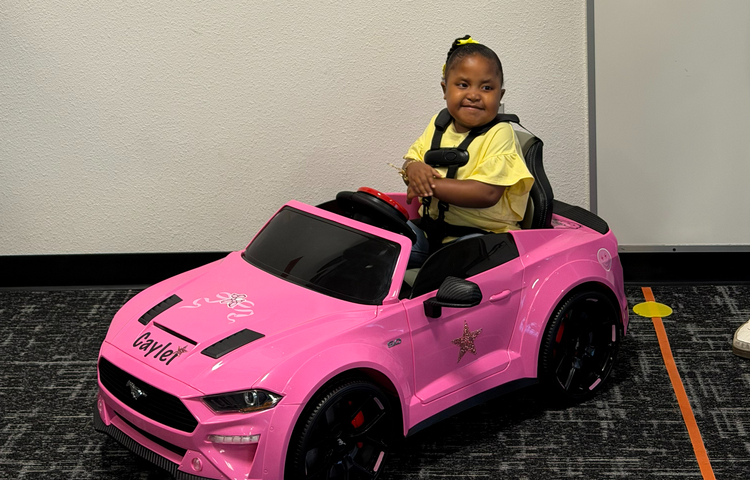 Little girl sitting in pink car with balloons and a sign that says Go Caylei Go