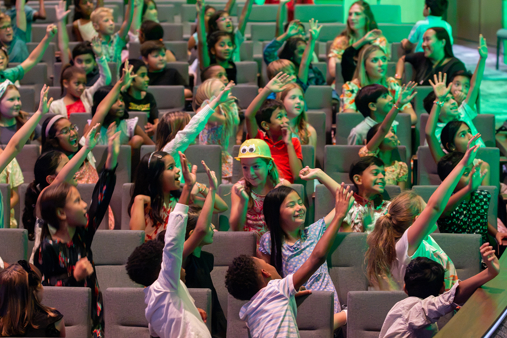 Students raise their hands to receive a duck hat handed out by the performers.