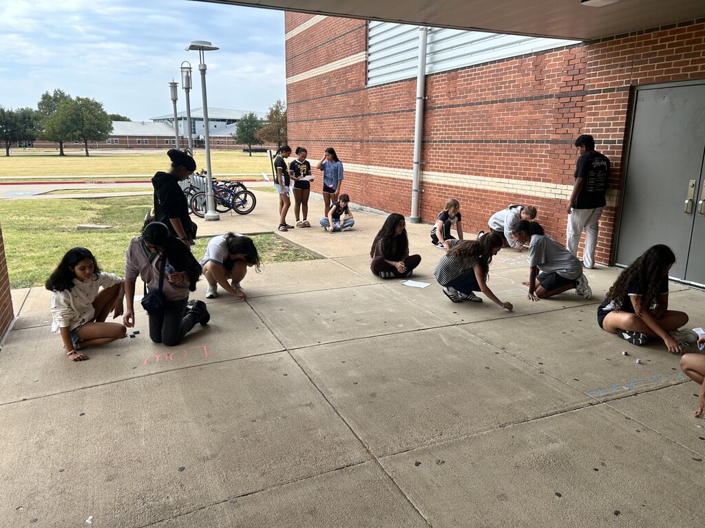 Yearbook staff members spread out along a school sidewalk, creating colorful chalk drawings and words that contain clues about this year’s yearbook theme. Students sit or kneel on the pavement working on their designs, while others stand nearby.
