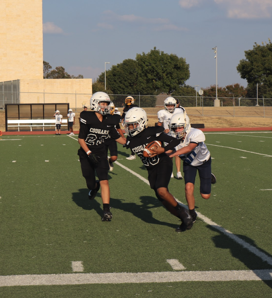 A Cougars football player carries the ball while an opposing player attempts to tackle him and a teammate runs alongside the play.