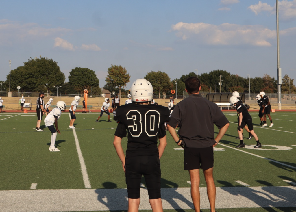 Football players from opposing teams line up at the line of scrimmage while coaches and teammates watch from the sideline.