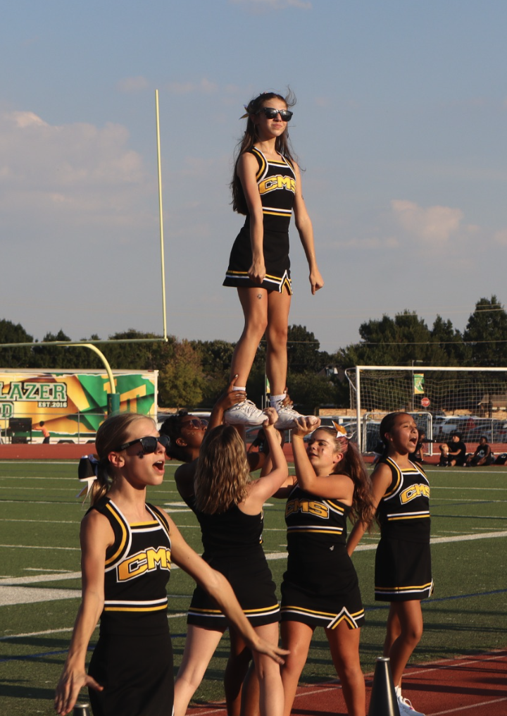 Clark Middle School cheerleaders perform a stunt on the sideline, lifting a teammate into the air during a football game.