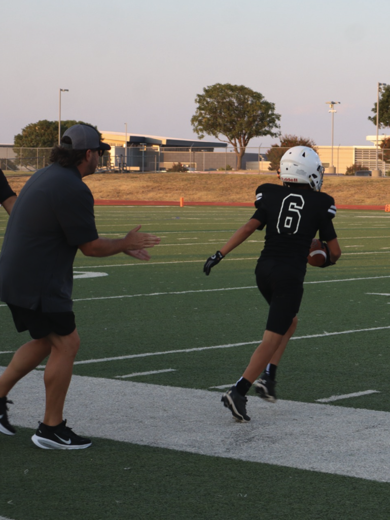 A Cougars football player carrying the ball runs down the sideline as a coach cheers him on.