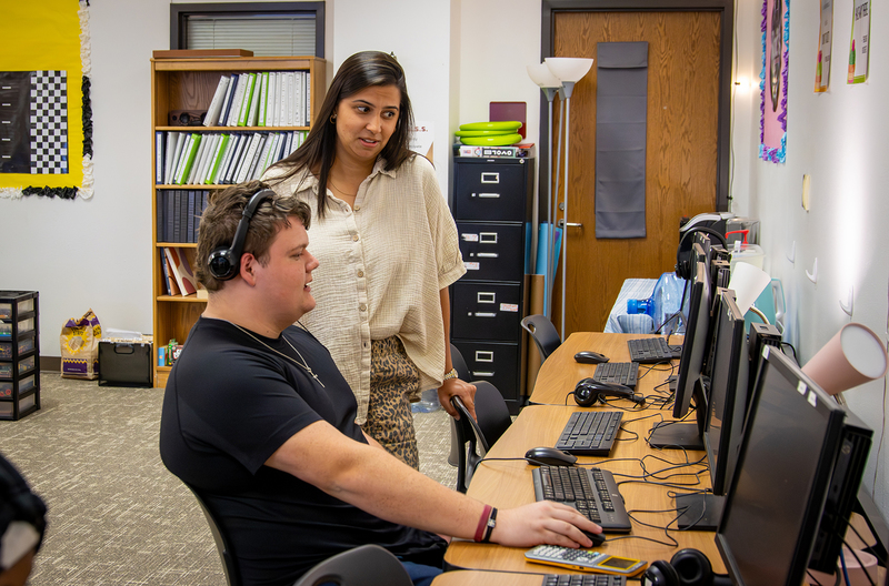 A student working on a computer and a teacher standing next to him and talking