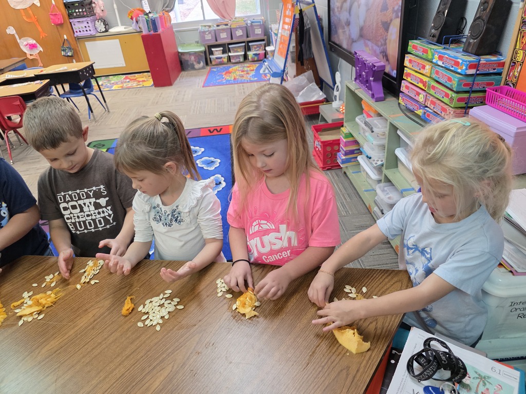 Four children at a table, one holding a banana, one holding a pumpkin, and one peeling a banana.