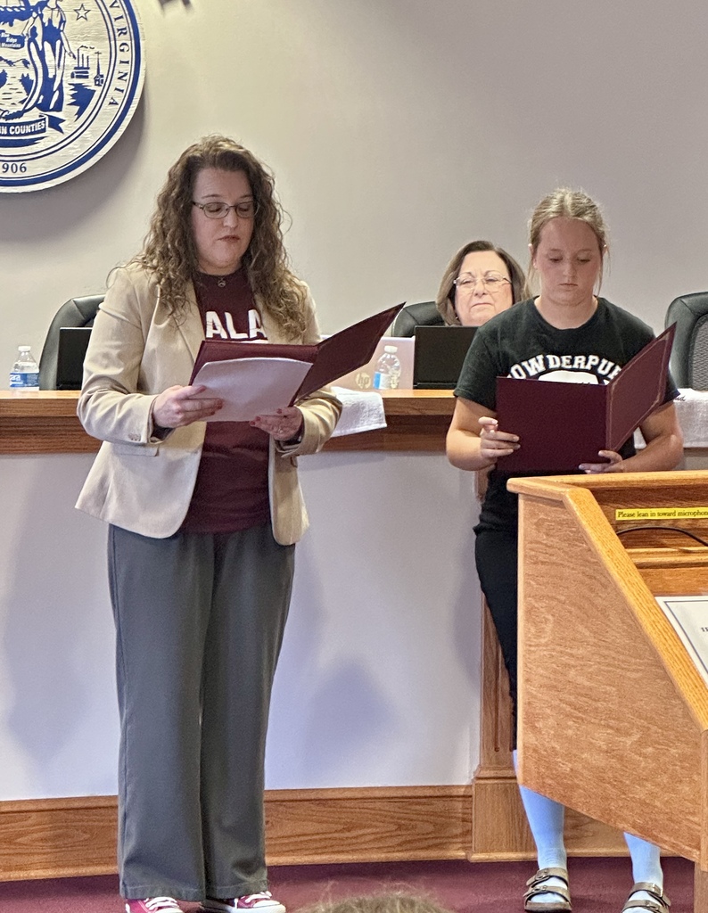 Two women hold open books, standing in front of a wooden podium with a microphone.