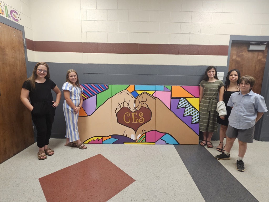 Six children pose next to a colorful mural of two hands with a heart on a wall.