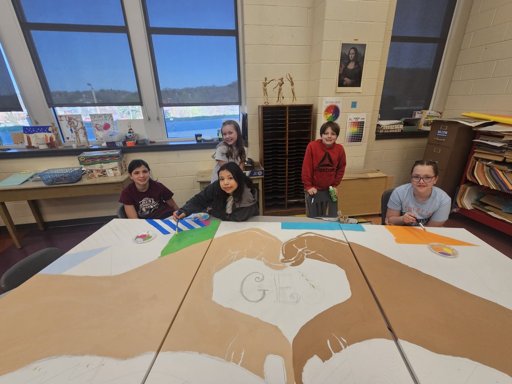 Five children in a classroom, gathered around a long table covered in a large hand painting.