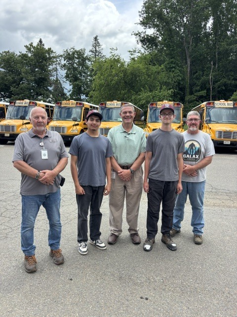 Five adult men stand in front of parked school buses, all wearing casual attire and standing on a paved surface.