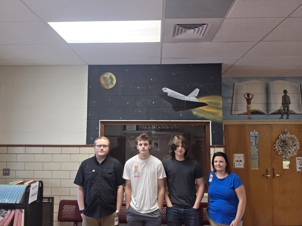 Four people stand in a room with a space shuttle mural and an open book on the wall.
