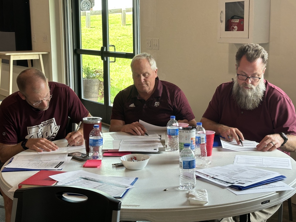 Four men sit around a table with papers, pens, and water bottles. They are in a room with a glass door.
