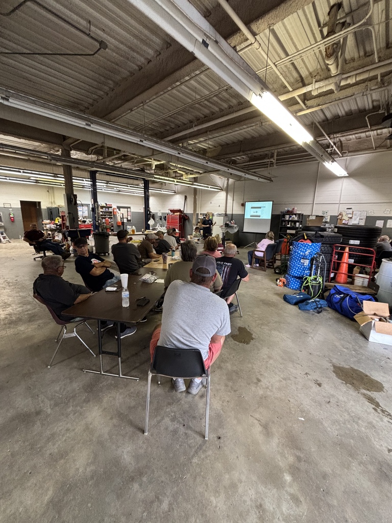 Group of people sitting in chairs around a table in a warehouse. A projector screen is visible on the wall.