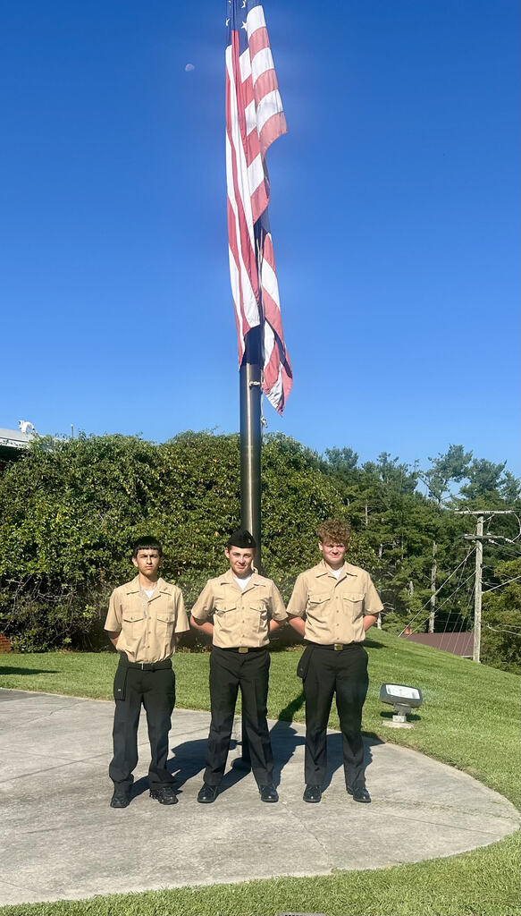 Three uniformed individuals stand before a flagpole with an American flag. They face forward with a green grassy background and trees.