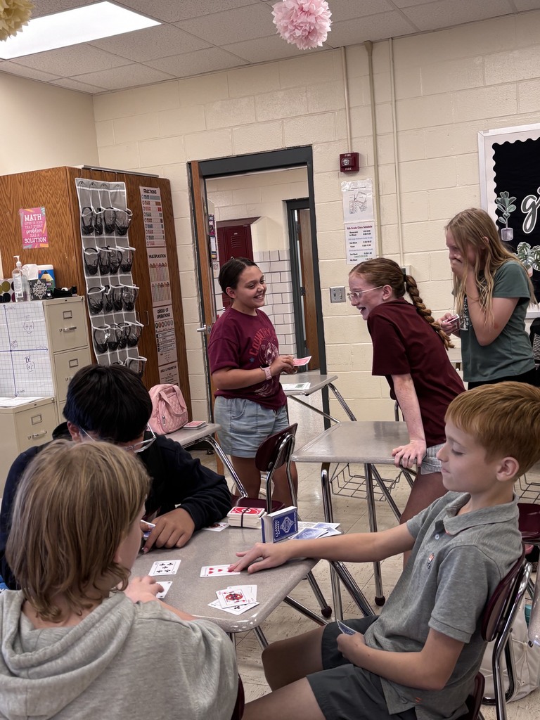 Six children seated around a table, playing cards in a classroom. Two women stand behind them, one gesturing.