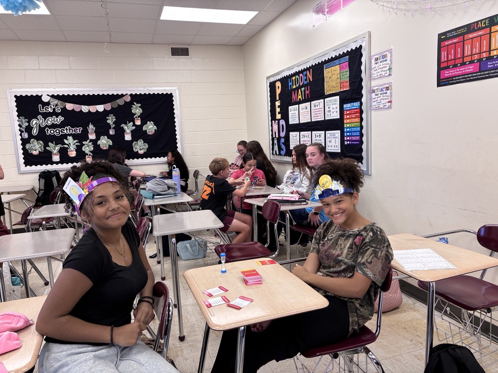 Students sit at desks in a classroom with a white wall. Some students wear headbands. Desks have papers, books, and a backpack.