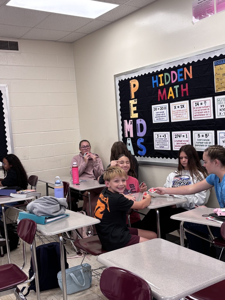 A classroom with students seated at desks. A board displays "HIDDEN MATH PEMDAS". A child holds a game.