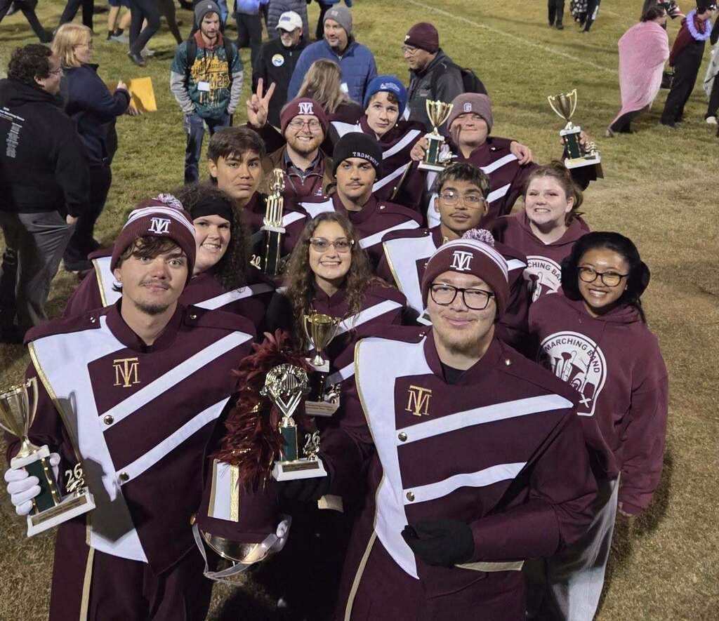 A group of students in maroon and white uniforms pose for a photo with trophies on a field.