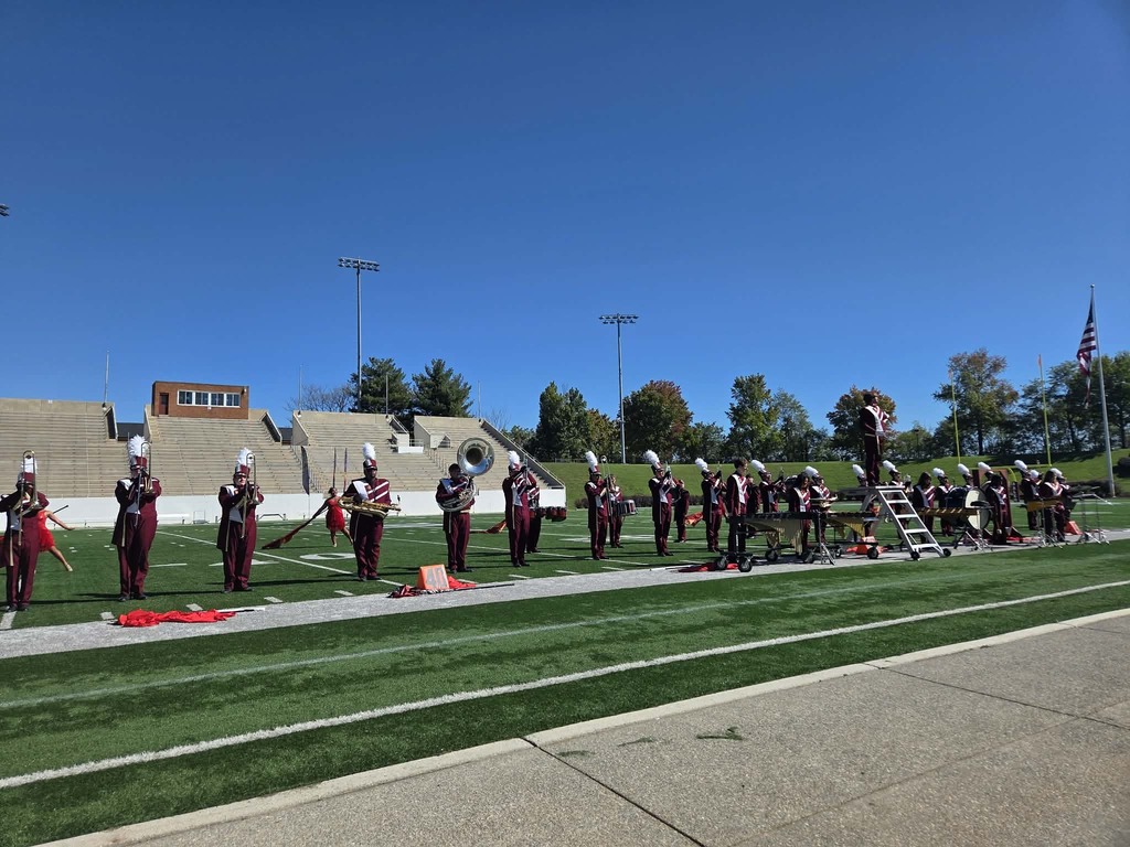 A band in red and black uniforms stands on a field, some holding instruments, with a clear sky in the background.
