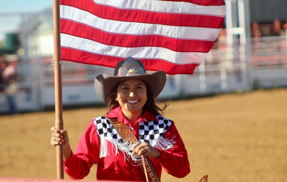 a young girl wearing a cowgirl hat, holding the american flag, smiling for a picture. She is on a horse. She is wearing red