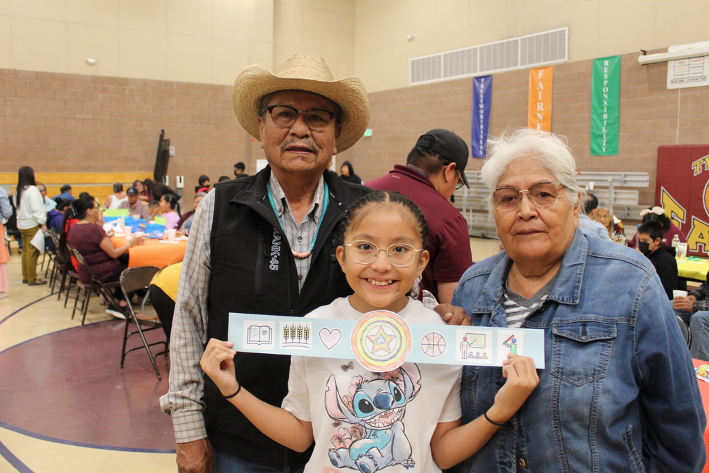 A grandmother and grandfather with their granddaughter. The grandchild is holding up a belt that she made with her grandparents.