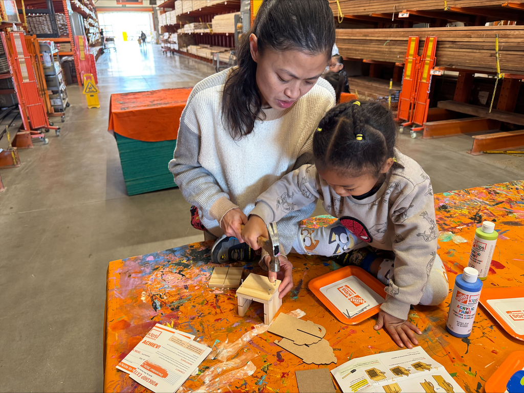 A woman and a child are indoors, using tools to assemble a small wooden structure on a table.