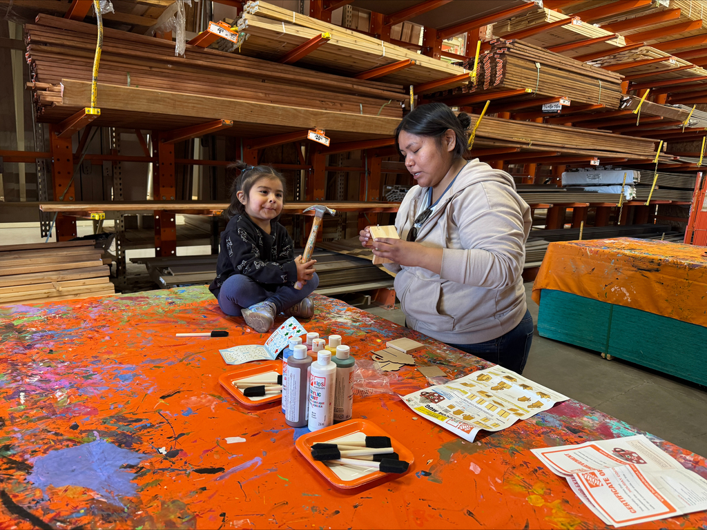 A woman and a child sit at a table, crafting with paint and brushes. Wooden shelves in the background.