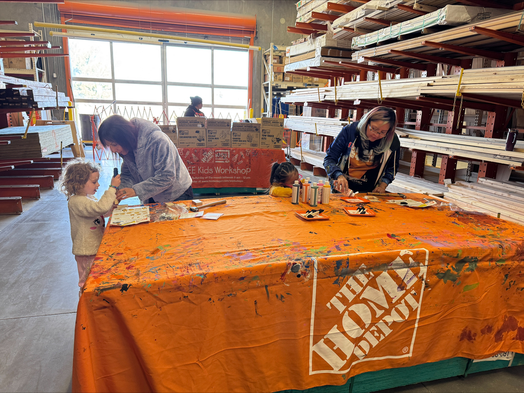 Three individuals, including two adults and a child, paint on an orange tablecloth at a Home Depot.
