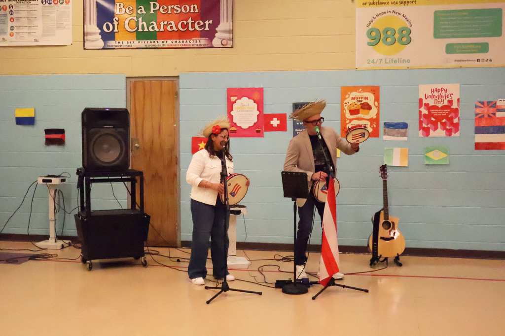 Two people perform on stage in a room. One holds a flag, while both hold a hand drum.