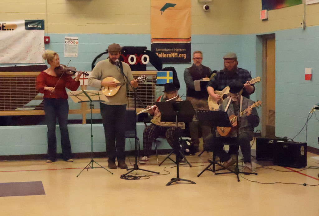 Six individuals are on stage with various musical instruments. Two play guitar, one fiddles, and one plays a banjo.