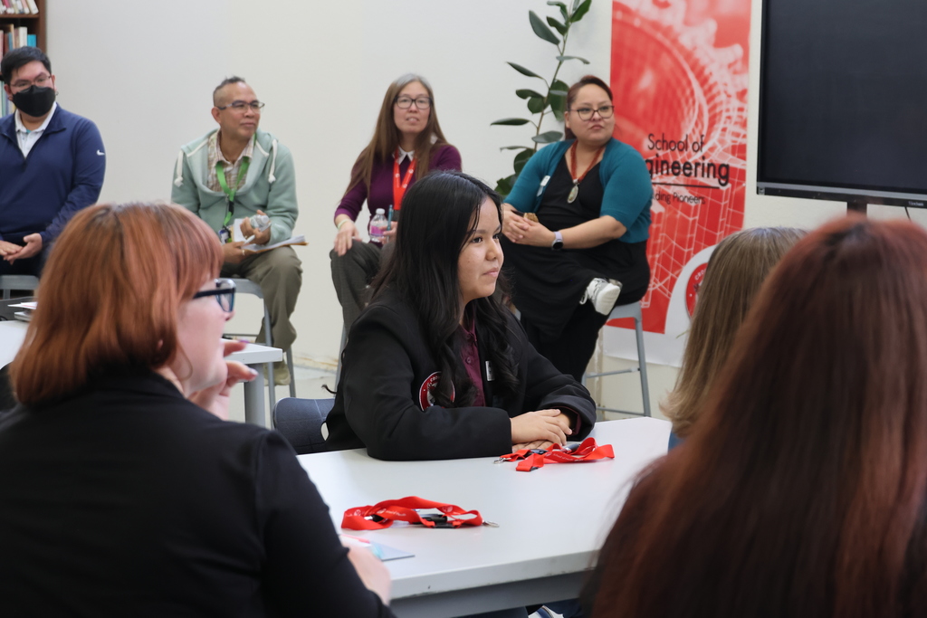 Several people sit around a table, engaged in conversation. One person speaks while others listen. Background features a red banner and a potted plant.