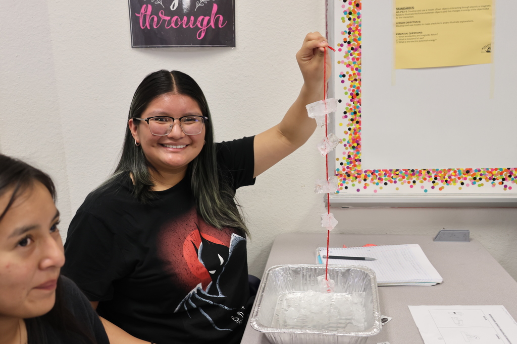 A woman in glasses and a Batman shirt stands and holds a string of beads. Another woman sits nearby. Behind them, a white board with a colorful border.