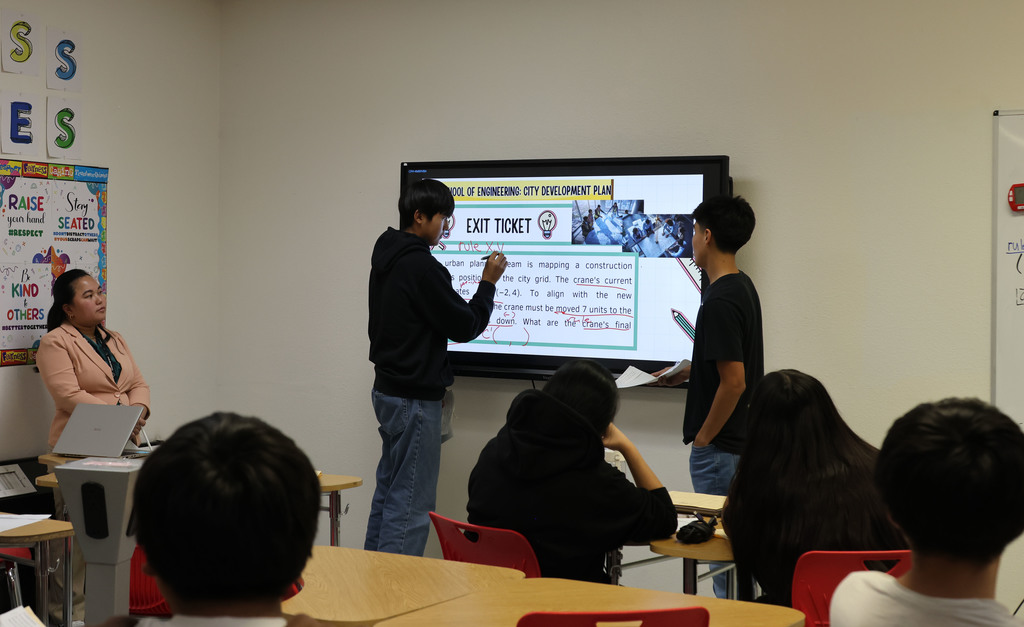 A classroom with a large screen displaying text. A person stands before the screen and writes. Students sit at desks.