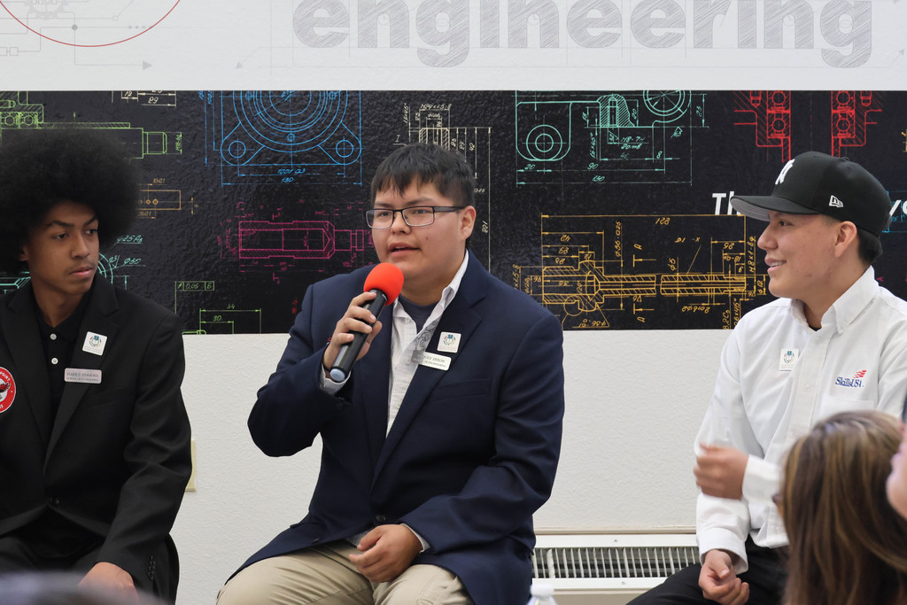 Three men seated in front of a colorful wall, one with a microphone speaking, others listening.